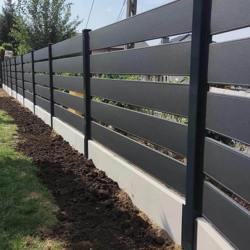 Nowoczesne ogrodzenie z poziomymi ciemnoszarymi panelami i czarnymi metalowymi słupkami stoi na niskiej betonowej podstawie na Woli Duchackiej, ze świeżo obróconą ziemią wzdłuż krawędzi i trawą po jednej stronie. W tle widoczne są drzewa i słup energetyczny.
