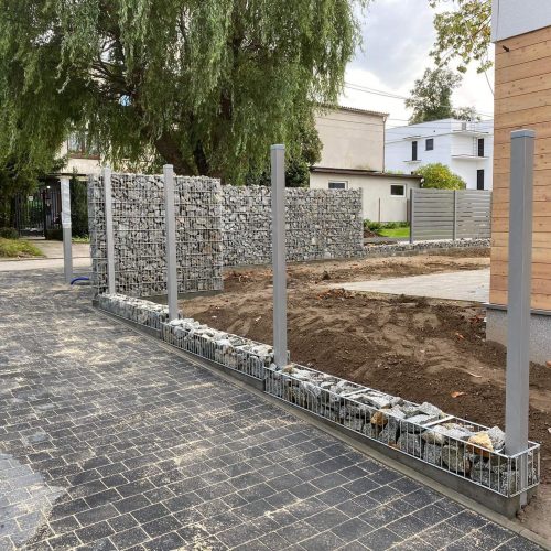 Częściowo zbudowane ogrodzenie gabionowe z metalowymi słupkami stoi obok wybrukowanego podjazdu i skrawka gołej ziemi, w pobliżu nowoczesnych domów i drzew liściastych w dzielnicy mieszkalnej.