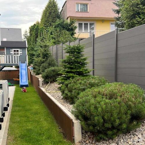 Wąskie podwórko z trawą, rzędem klimatyzatorów po lewej, krzewami i małymi drzewami pośrodku oraz szarym ogrodzeniem po prawej. W tle widoczna jest niebieska zjeżdżalnia i dom.