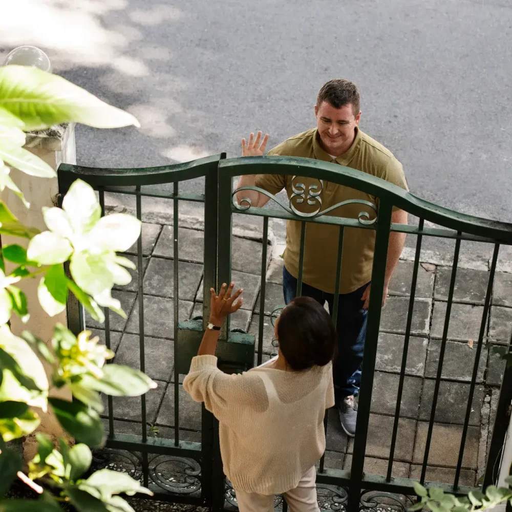 Mężczyzna stoi przed metalową bramą, uśmiechając się i machając do kobiety po drugiej stronie, która sięga w kierunku bramy. Słoneczna, zielona scena sugeruje niedawny montaż ogrodzeń Kraków lub przygotowanie działki pod ogrodzenie.