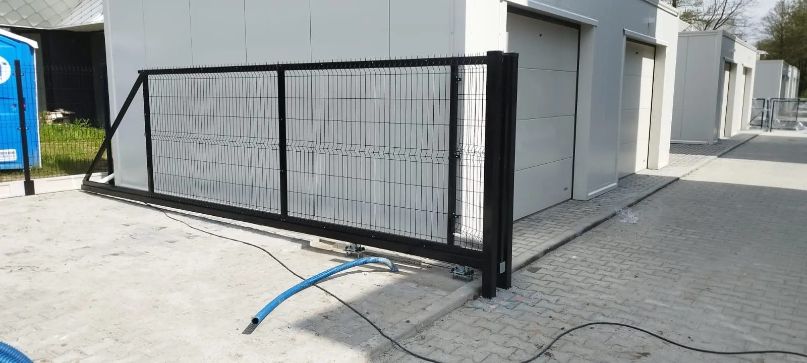 Czarna metalowa brama przesuwna zainstalowana na wybrukowanym podjeździe obok nowoczesnego białego budynku przemysłowego, z kablami i niebieskim wężem leżącymi na ziemi.