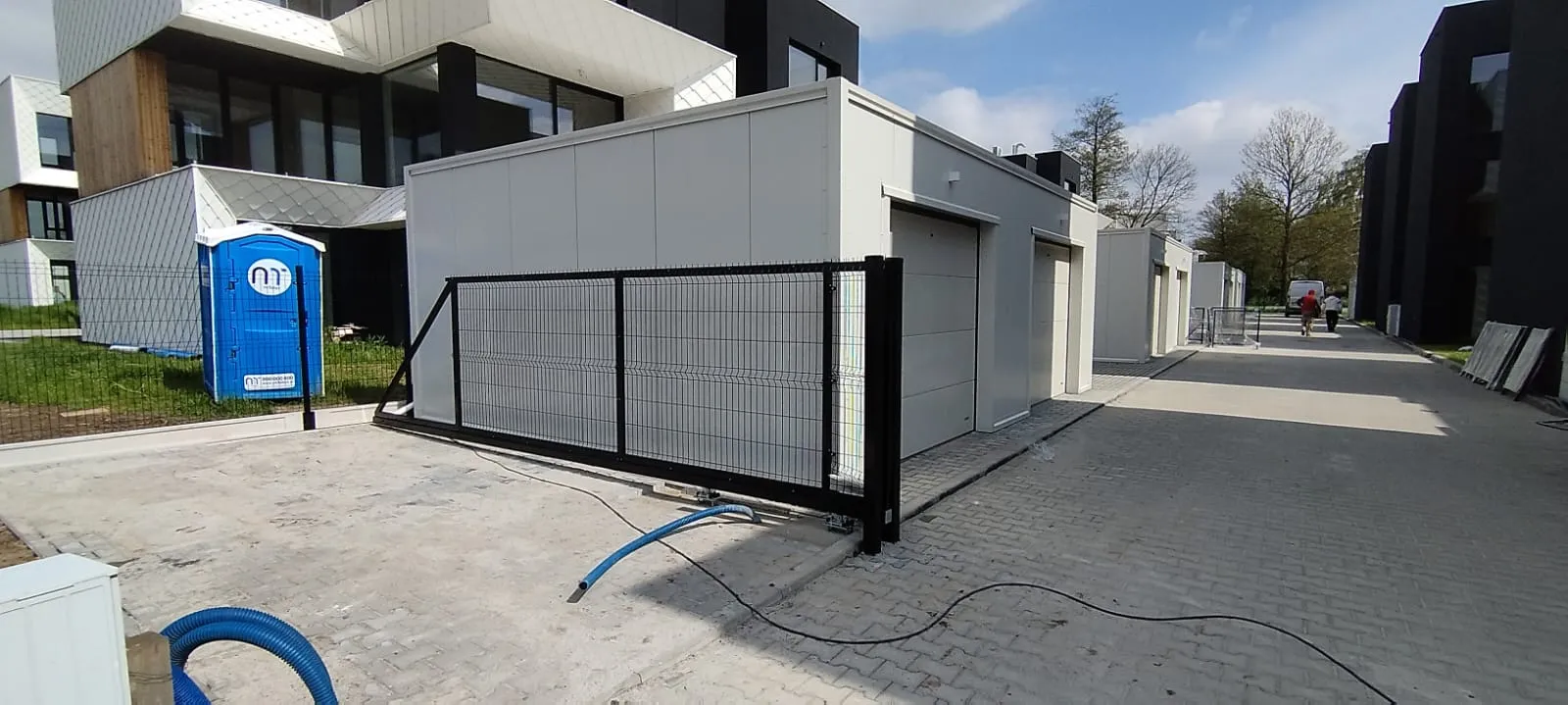 Nowoczesna dzielnica mieszkaniowa z białymi budynkami modułowymi, przenośną toaletą i czarną metalową bramą przesuwną częściowo otwartą na wybrukowanym podjeździe. Na ziemi leżą niebieskie kable. W tle widoczne są drzewa.