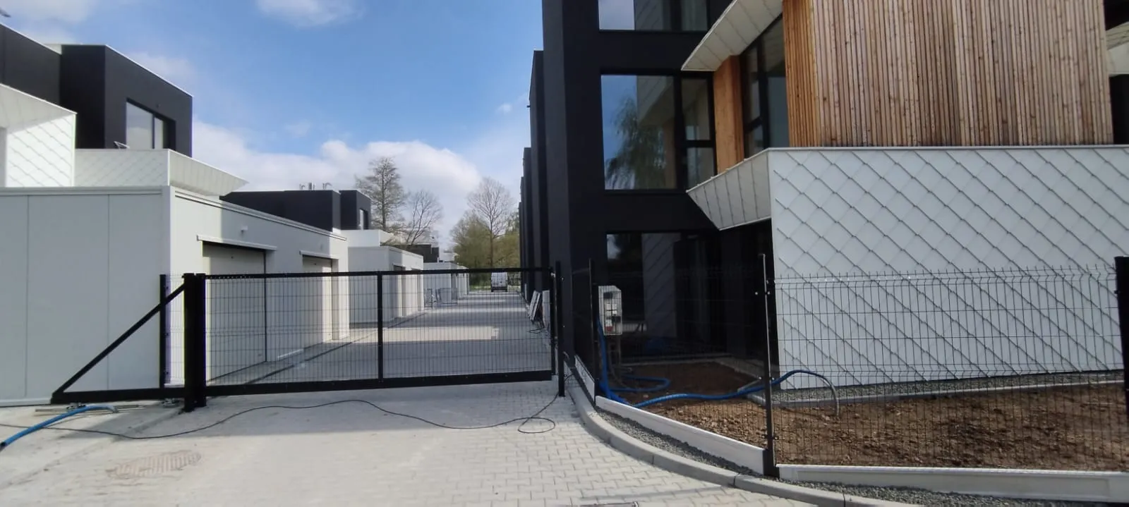 Nowoczesne osiedle zamknięte z nowoczesnymi budynkami, czarnym metalowym ogrodzeniem, zamkniętą bramą, wybrukowanym podjazdem i gołą ziemią wzdłuż chodnika. Niebo jest częściowo zachmurzone.