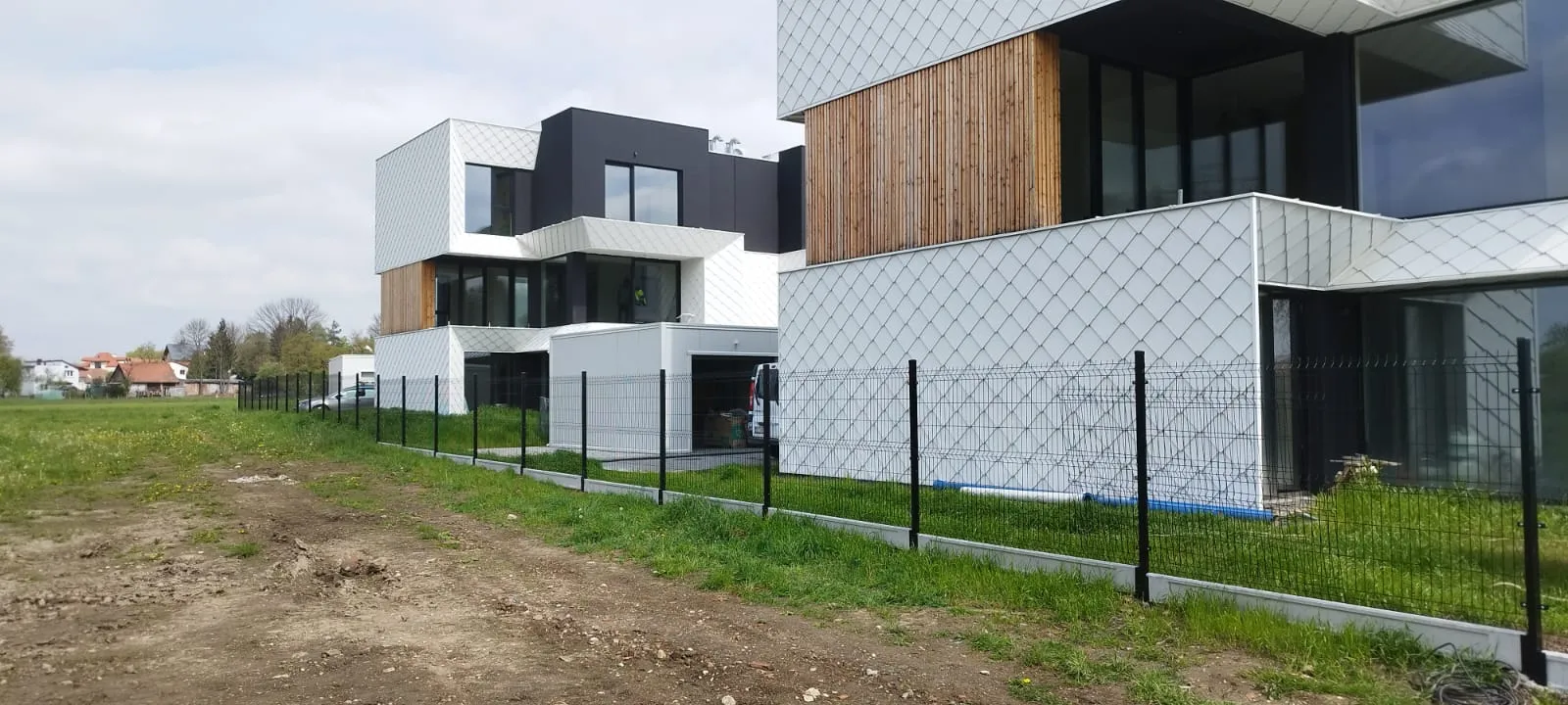 Nowoczesne, geometryczne budynki mieszkalne z białymi i drewnianymi elewacjami są oddzielone od trawiastego, otwartego terenu czarnym metalowym ogrodzeniem. Ścieżka biegnie wzdłuż ogrodzenia; domy i drzewa widoczne w tle.