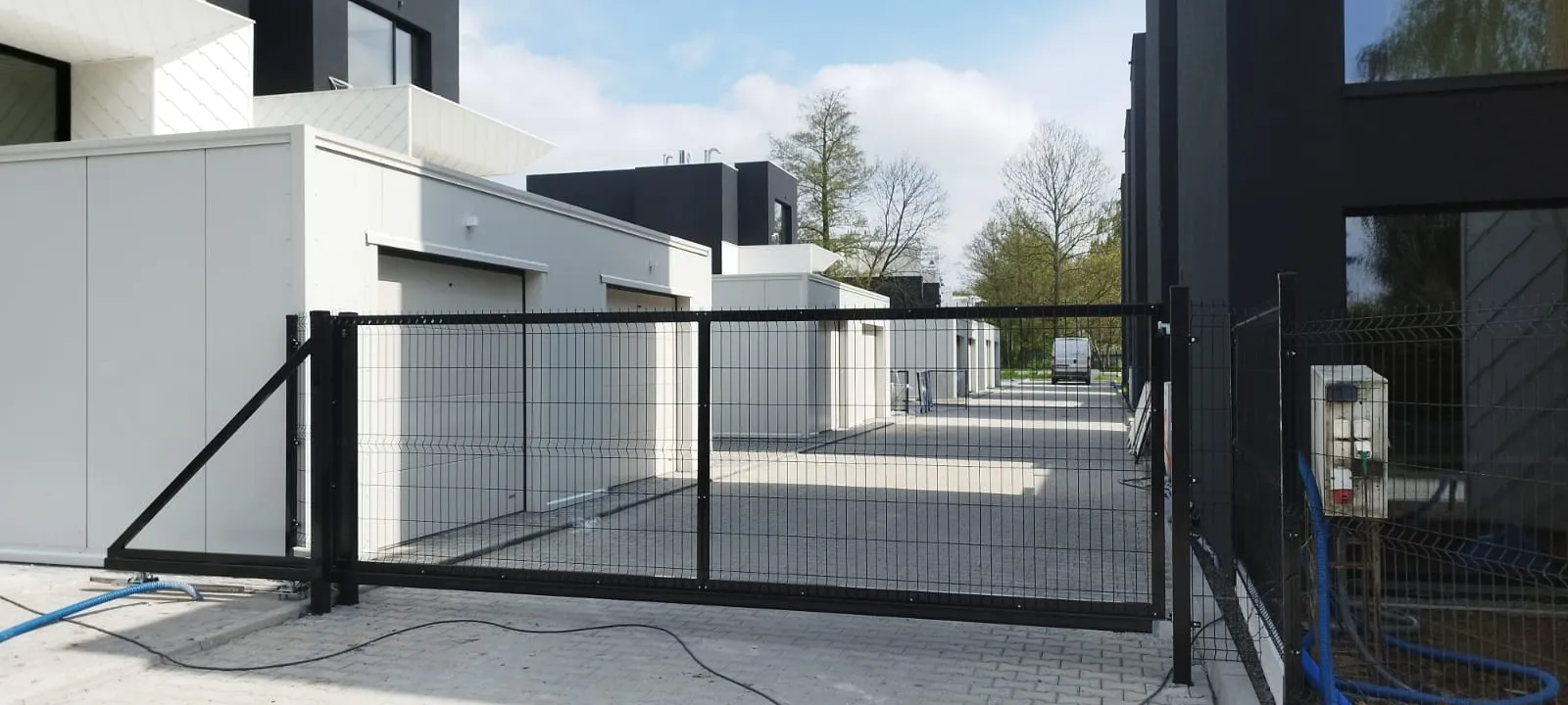 Nowoczesna czarna brama przesuwna blokuje wjazd do kompleksu mieszkalnego z białymi i ciemnoszarymi budynkami, wybrukowanym podjazdem i kilkoma drzewami widocznymi w tle pod częściowo zachmurzonym niebem.