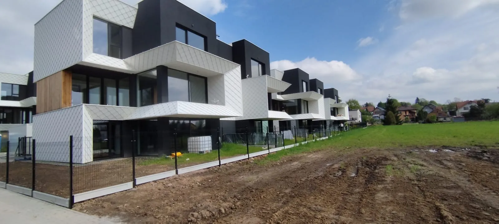 Nowoczesny rząd kanciastych białych i czarnych kamienic z dużymi oknami i balkonami, oddzielonych czarnymi płotami, obok trawiastego i błotnistego otwartego terenu pod częściowo zachmurzonym niebem.