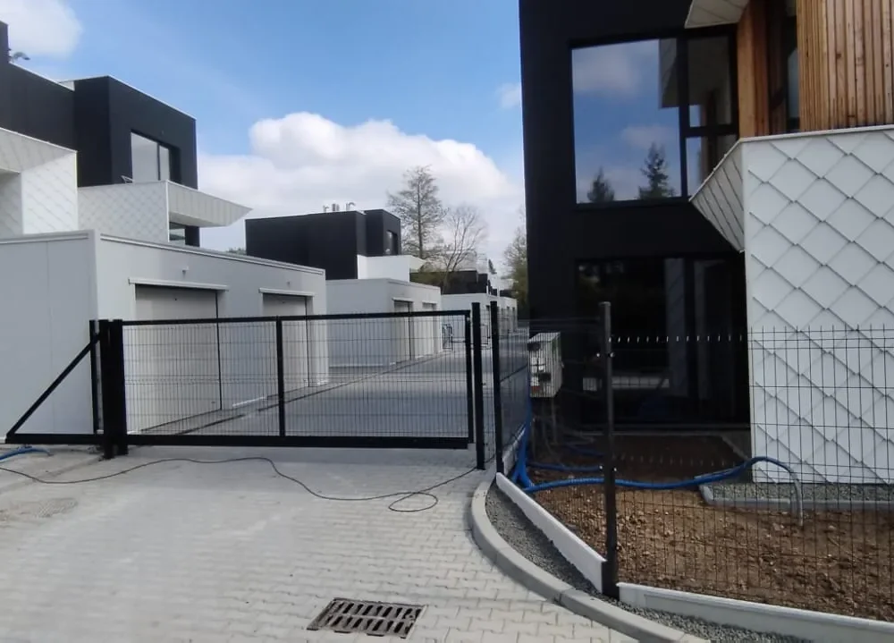 Nowoczesny kompleks mieszkalny z białymi i czarnymi domami przypominającymi kostkę, garażami, wybrukowanym podjazdem i czarną metalową bramą na pierwszym planie. Obszar wygląda na czysty i nowo wybudowany, z niedokończonym zagospodarowaniem terenu.