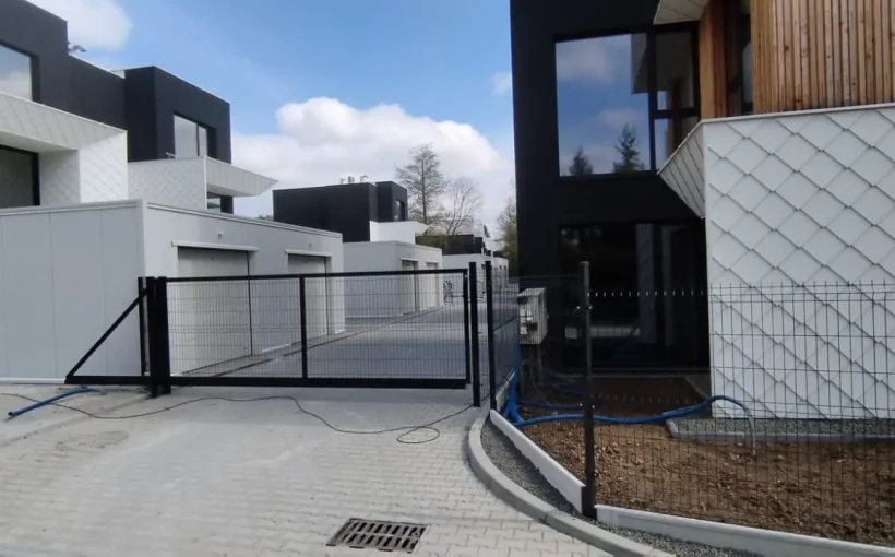 Nowoczesny kompleks mieszkalny z białymi i czarnymi domami przypominającymi kostkę, garażami, wybrukowanym podjazdem i czarną metalową bramą na pierwszym planie. Obszar wygląda na czysty i nowo wybudowany, z niedokończonym zagospodarowaniem terenu.