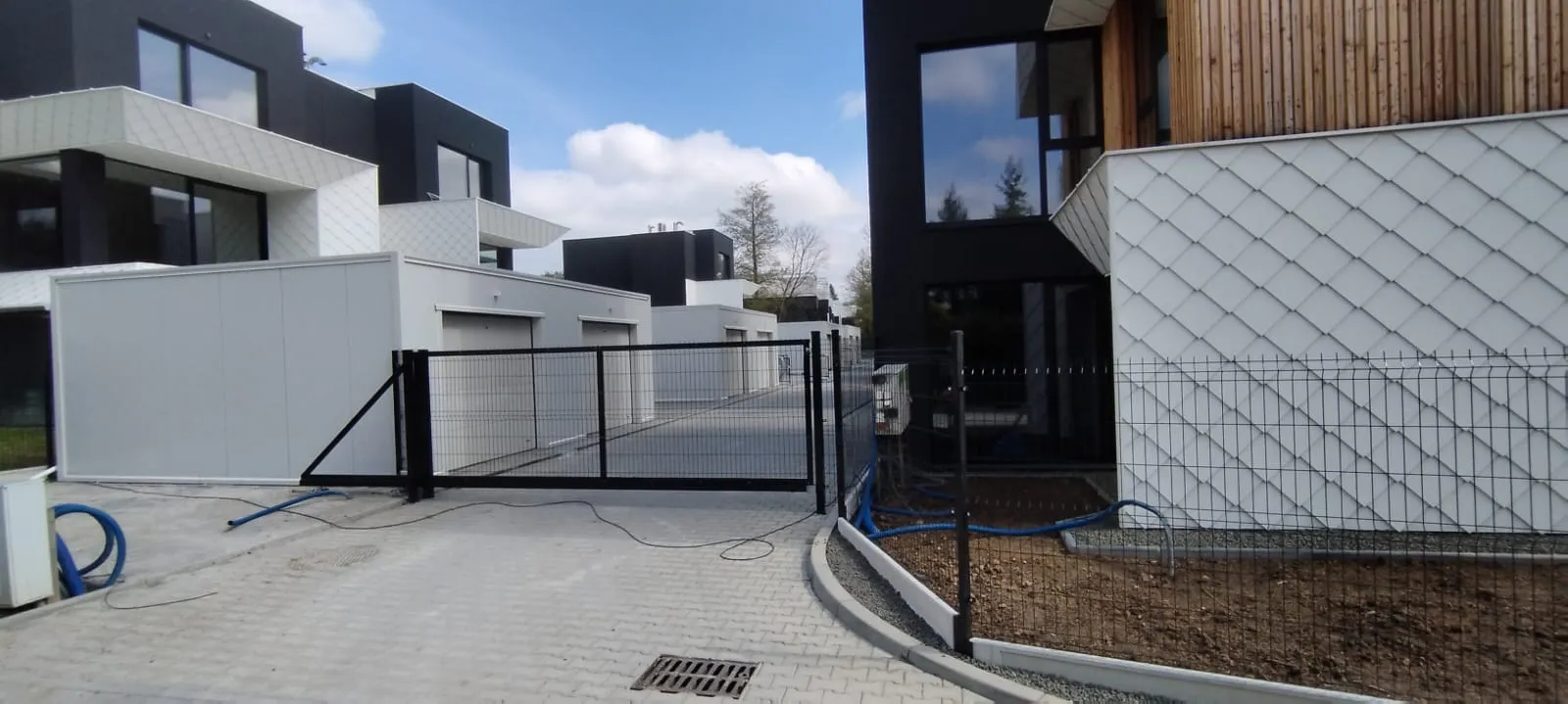 Nowoczesny kompleks mieszkalny z białymi i czarnymi domami przypominającymi kostkę, garażami, wybrukowanym podjazdem i czarną metalową bramą na pierwszym planie. Obszar wygląda na czysty i nowo wybudowany, z niedokończonym zagospodarowaniem terenu.
