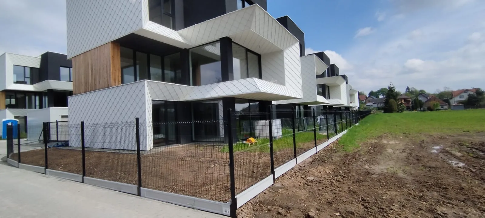 Nowoczesne, kanciaste budynki mieszkalne z dużymi szklanymi oknami i białymi geometrycznymi fasadami stoją za czarnym metalowym ogrodzeniem obok skrawka ziemi i trawiastego pola pod częściowo zachmurzonym niebem.