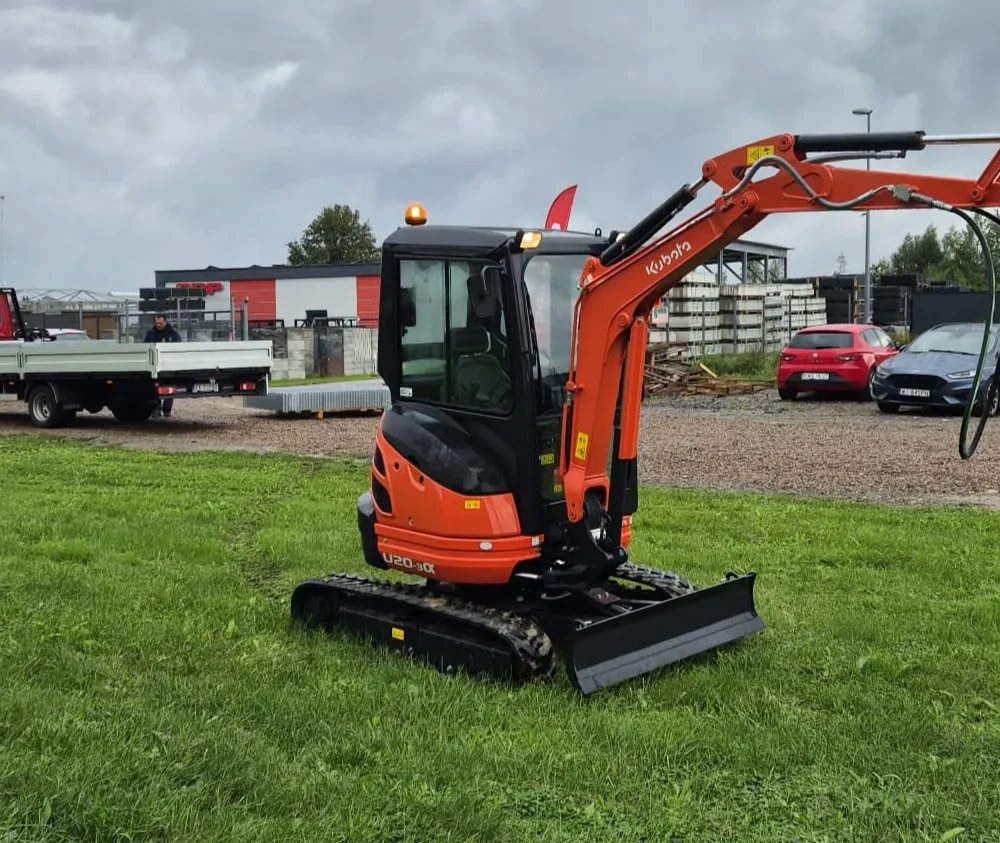 Pomarańczowa minikoparka Kraków z osprzętem ślimakowym jest zaparkowana na trawiastym terenie. Za nią znajduje się kilka pojazdów, w tym czerwony samochód i ciężarówka z platformą, a w tle budynki pod zachmurzonym niebem.