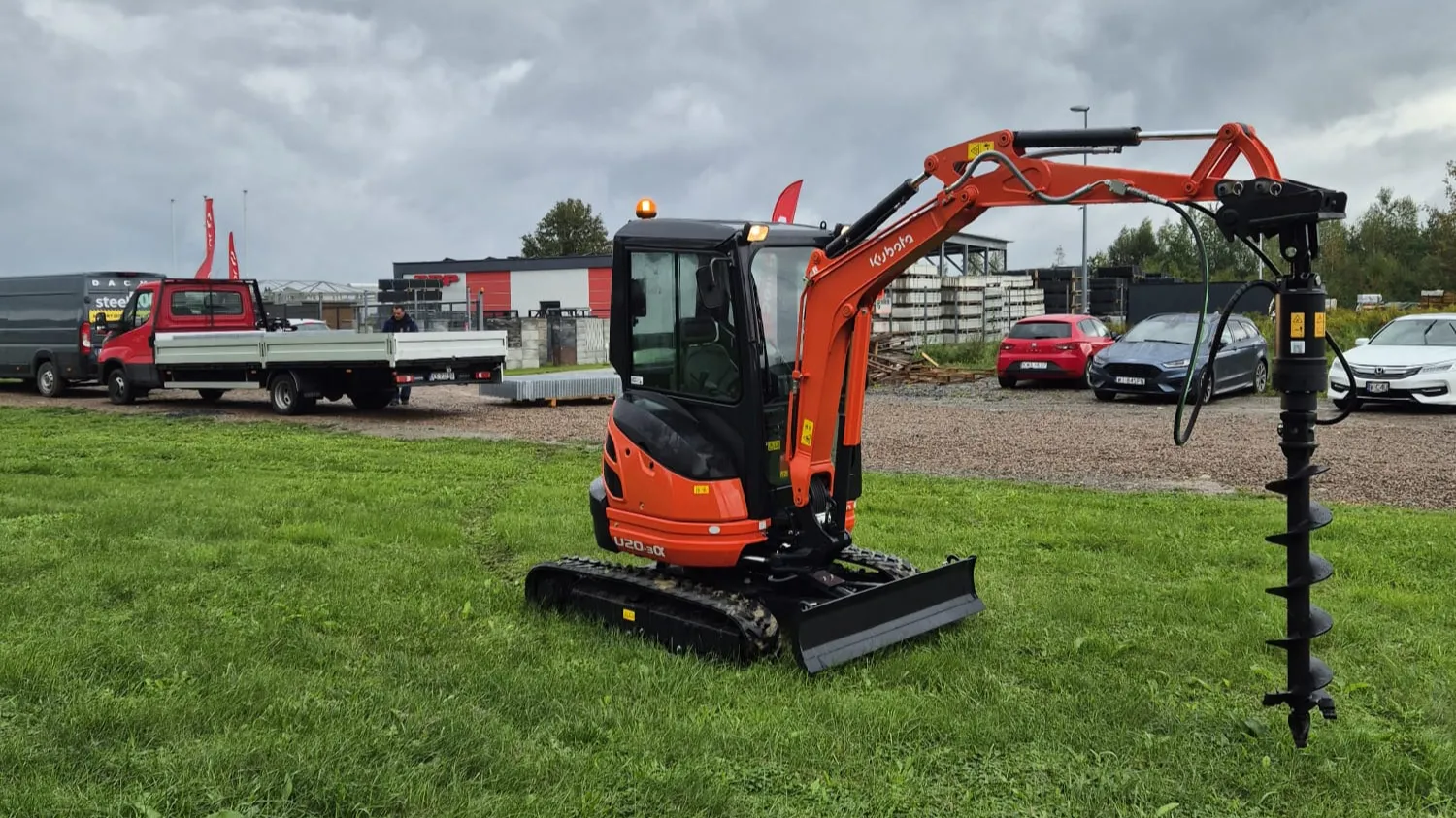 Pomarańczowa minikoparka Kraków z osprzętem ślimakowym jest zaparkowana na trawiastym terenie. Za nią znajduje się kilka pojazdów, w tym czerwony samochód i ciężarówka z platformą, a w tle budynki pod zachmurzonym niebem.