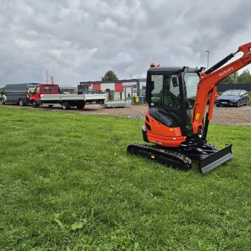 Mała pomarańczowa koparka, idealna do koparka wynajem Małopolska, jest zaparkowana na zielonej trawie pod zachmurzonym niebem. W tle widać czerwoną lawetę i budynek ze znakiem Scania.