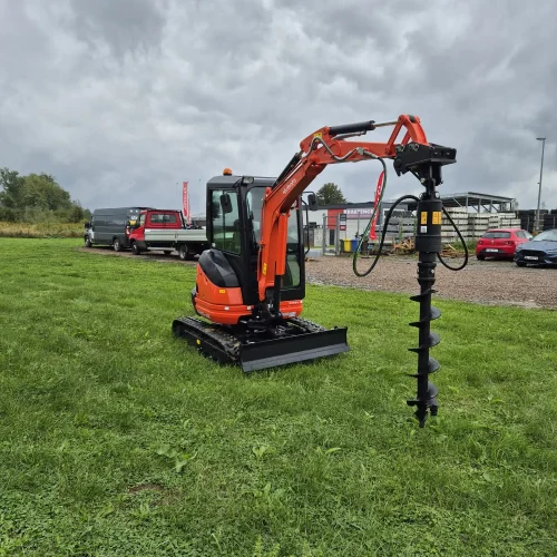 Pomarańczowa minikoparka z osprzętem ślimakowym, idealna do kopania pod fundamenty ogrodzenia, jest zaparkowana na zielonej trawie pod zachmurzonym niebem. W tle widać samochody i budynki - koparka wynajem Małopolska dostępna.