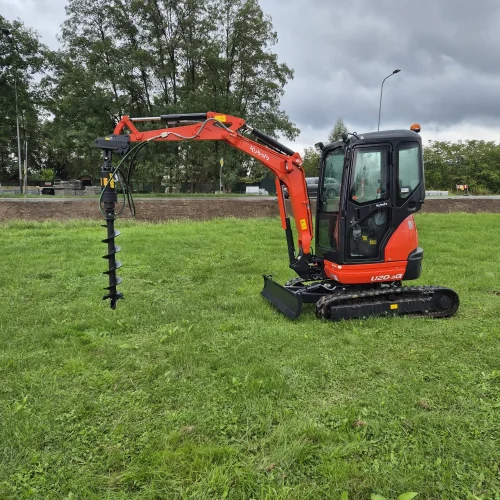 Mała pomarańczowa koparka z osprzętem ślimakowym jest zaparkowana na trawiastym polu pod zachmurzonym niebem, gotowa do otworów pod ogrodzenia. W tle widoczne są drzewa i droga z samochodami.